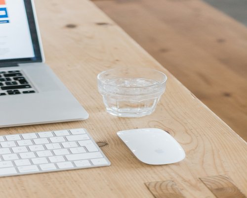 A laptop on an organized wooden desk surface next to a glass of water and natural light filters